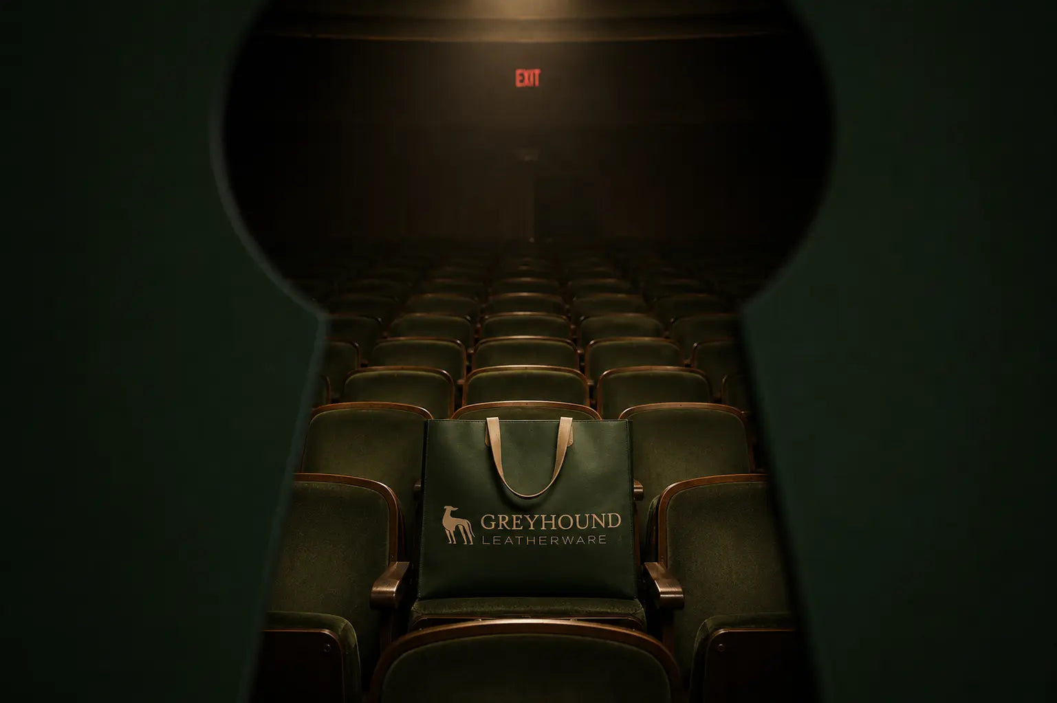 A dark green tote bag with the Greyhound Leatherware logo sits in the center of an empty movie theater row of seats, viewed through a keyhole-shaped frame looking out toward the stage.