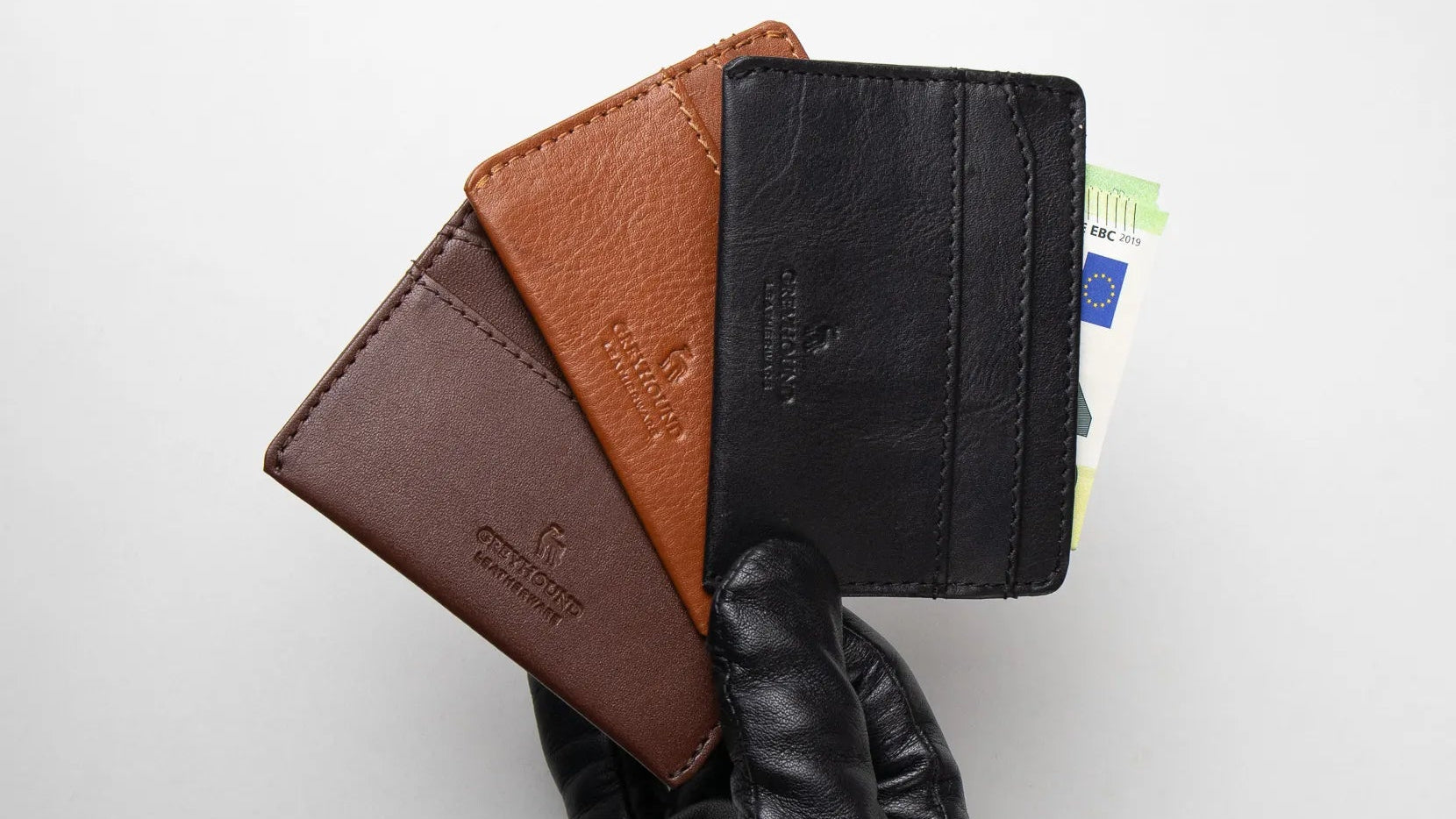 Three leather wallets in brown, black, and tan held by a gloved hand on a white background.